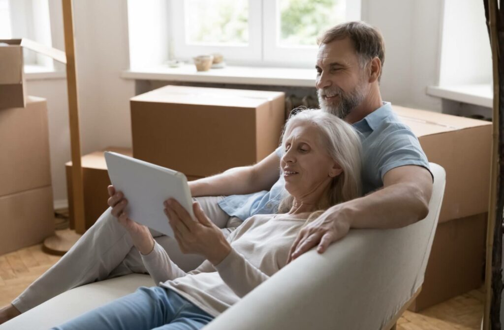 A senior couple cuddle together on a couch, surrounded by moving boxes, researching retirement communities on a tablet