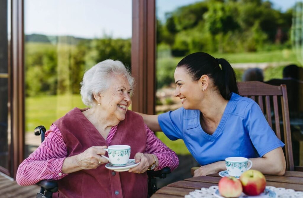 A caregiver and an older adults mile at one another while sitting in a sunroom and drinking tree in front of a beautiful green space