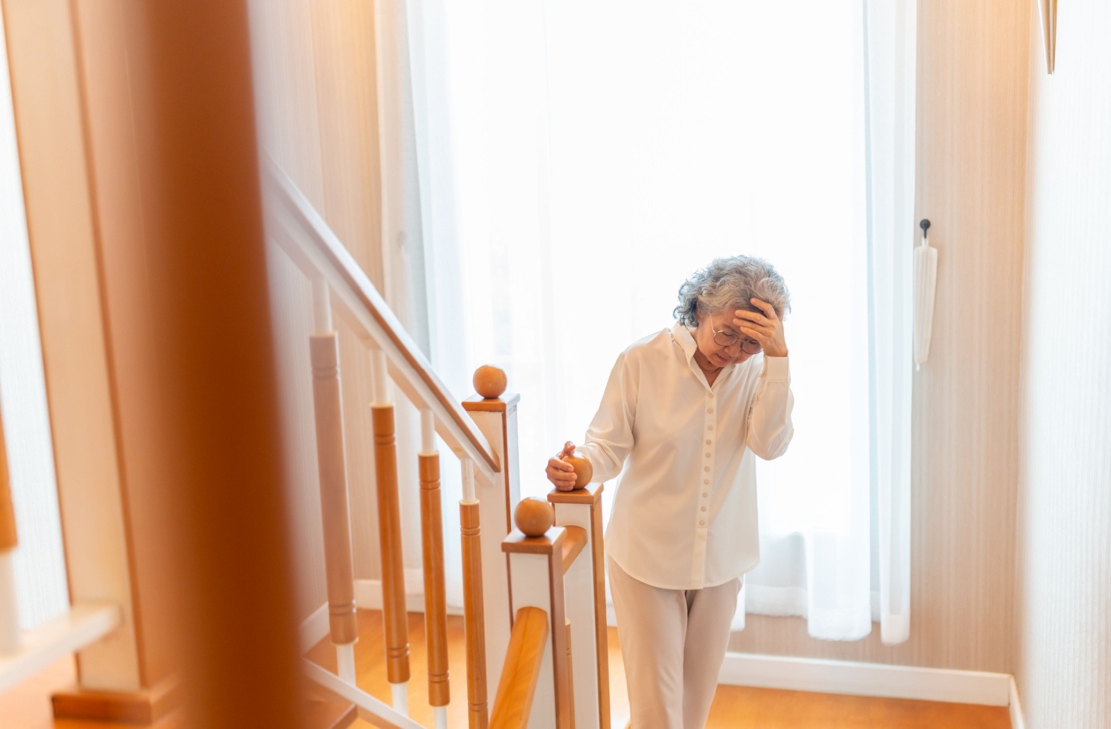 A frustrated senior holds their head at the bottom of the stairs in their home.